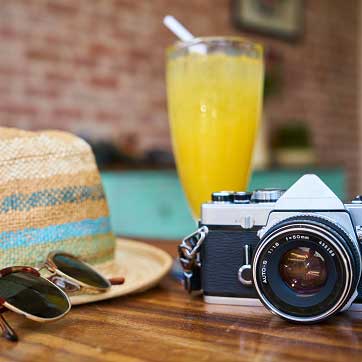 Billedet forestiller et par solbriller, et kamera, en solhat og et glas juice på et bord.