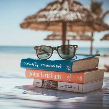 Billedet viser nogle bøger og et par solbriller, som ligger på et bord på en strand.